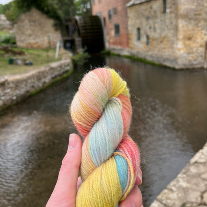 A white hand holds a skein of rainbow yarn in front of a picturesque old water mill.